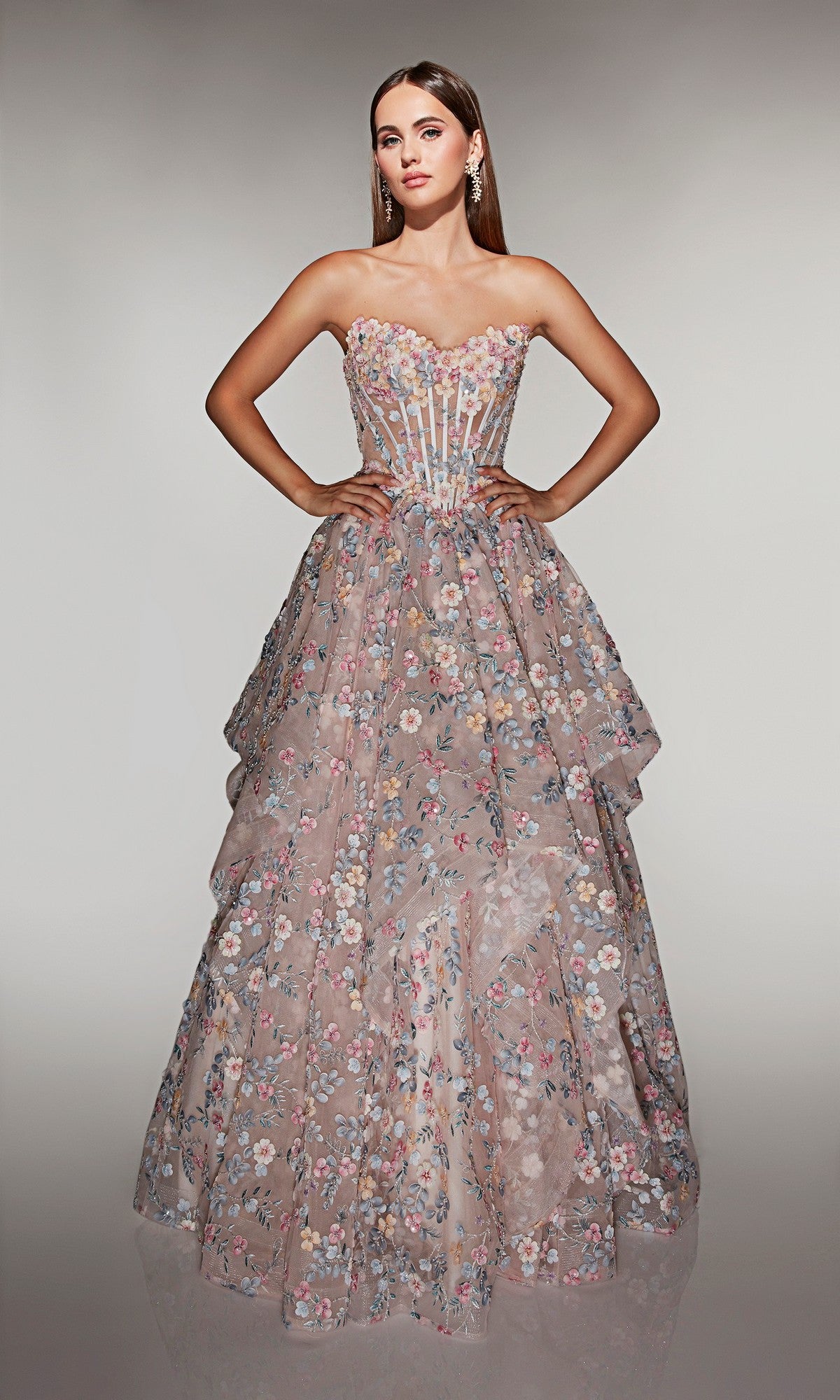 A woman stands against a neutral background wearing the Alyce 62048 long prom dress in French Pink, featuring a strapless fitted corset bodice, layered skirt, pastel floral pattern, and sheer details.